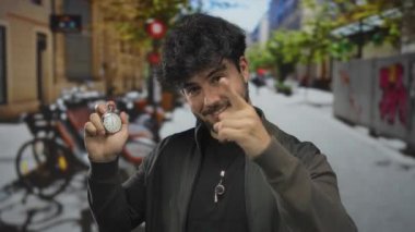 Sakallı, kronometre tutan ve güneşli caddede parmak gösteren, arka planda bulanık bisiklet ve binaları olan genç İspanyol bir adam, aciliyet ve eylem öneriyor..