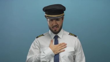 Sakallı ve üniformalı genç pilot, kafa karışıklığını ifade eden, havayolu, İspanyol ve profesyonel havacılık temalarını vurgulayan mavi bir arka plana karşı duruyor..