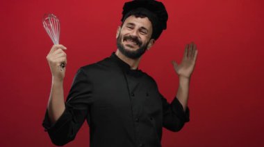 Sakallı İspanyol erkek aşçı neşeli bir şekilde çırpıcıyı canlı kırmızı arka planda tutuyor, aşçılık hevesini ve profesyonel kıyafetlerini sergiliyor..