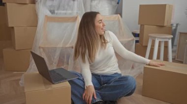 Genç kadın yeni evdeki kutuların arasında oturmuş dizüstü bilgisayarla gülümsüyor ve modern iç dekorasyonda paketleri açıyor..