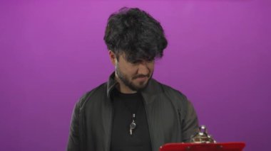 Sakallı genç adam pembe arka plana yaslanmış elinde mor bir arkaplan olan bir pano tutuyor..