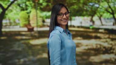 Canlı yeşil bir park ortamında parmaklarını gösteren genç bir kadın, gözlük ve mavi gömlek takıyor, güneşli bir açık havada pozitifliğini ve güvenini ifade ediyor..