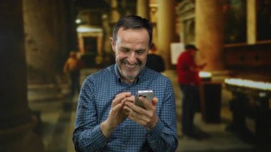 Kilisenin içinde arka planı bulanık ve sütun yapılarıyla gülümseyen bir adam samimi bir sevinç ve teknoloji etkileşimi yakalıyor..