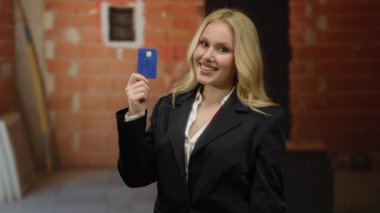 İnşaat alanında kredi kartı tutan bir kadın, siyah takım elbiseli, sarı saçlı, kendine güvenen, içten gülümseyen, tuğla duvarlar görünür, sergilenen finansal konsept, neşeli ifade..