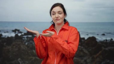Turuncu tulum giymiş bir kadın kayalık plaj arka planında el hareketi yapıyor..
