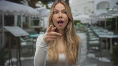 Kafkasyalı sarışın kadın restoranın terasında kamerayı işaret ediyor; sürpriz davetiye ile heyecan nişanı.