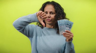 Orta yaşlı bir kadın Çin Yuan banknotlarını elinde tutarken, izole edilmiş sarı bir arkaplan üzerinde onaylanmamış veya mali tatminsizliği ifade eden baş parmağıyla bir jest yapıyor..