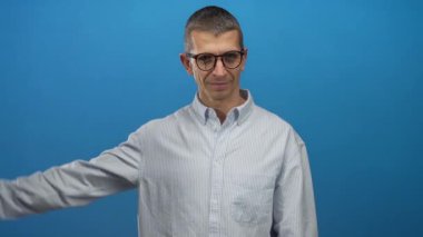 Gözlüklü beyaz adam, izole edilmiş mavi duvar arka planına karşı zafer jesti yapıyor, çizgili bir gömlek giyiyor, kendine güvenini ve başarısını iletiyor..