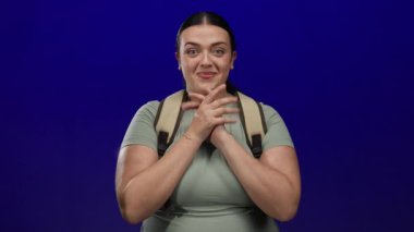 Mutluluğunu ifade eden genç bir kadın mavi arka plana karşı neşeli hareketlerle günlük kıyafetler ve sırt çantası giyiyor.