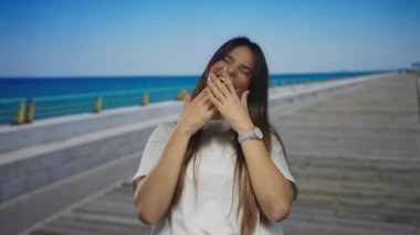 Deniz kenarındaki güneşli bir güvertede neşeyle gülen bir kadın. Dalgalarla dolu, günlük giysiler giymiş ve dalgalı okyanus ufkuna doğru ilerlerken bulaşıcı bir gülümseme takınmış..