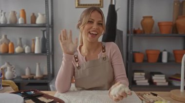 Çömlek atölyesinde kadın el sallarken ve önlük giyerken; dostluk yaratıcılığı ısıtır.