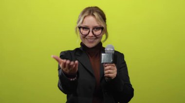 Elinde mikrofon tutan gözlüklü muhabir kadın canlı mavi arka planda şık bir ceket giyiyor..