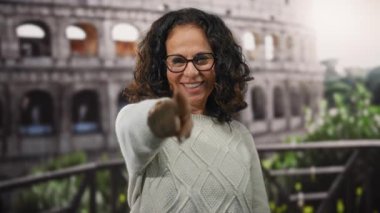 Kıvırcık saçlı olgun bir kadın Roma Kolezyumu önünde kalp hareketi yapıyor. Roma, İtalya 'da ikonik bir açık hava ortamında mutluluğu ve kültürü sergiliyor..