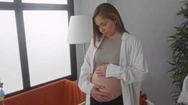 Hamile kadın düşünceli bir bebek odasında duruyor, göbeğini nazikçe kucaklıyor, yumuşak ışıklandırma, rahat bir beşik ve huzurlu bir iç dekorasyon ile çevrili..