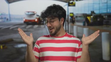 Sakallı genç İspanyol adam havaalanına şaşkın görünüyor. Çizgili tişörtü ve açık elleri olan, arka planda bir terminale ve arabalara karşı kurulmuş..