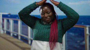 Gezgin gemisinde örülmüş saçlarıyla gülümseyen bir kadın okyanus manzarasına karşı üçgen oluşturuyor, açık havada neşe ve rahatlama yayıyor..