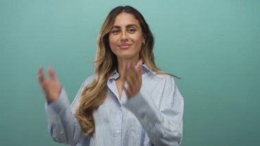 Mavi bir stüdyoda çıplak el sallayan ve gülümseyen uzun sarı saçlı ve çıplak yüzlü bir kadın, sevinç heyecanını selamlıyor..