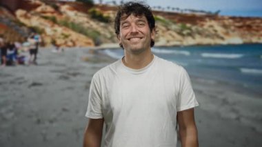 Beyaz tişörtlü İspanyol adam, güneşli bir plaj gününde neşeyle gülümsüyor, dalgalar arka planda çarpıyor, deniz kenarında mutluluk ve rahatlama ifade ediyor..