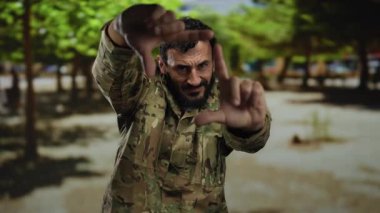 Askeri üniformalı İspanyol bir adam ağaçlarla dolu bir sokakta dikilirken manzarasını elleriyle çerçeveliyor..