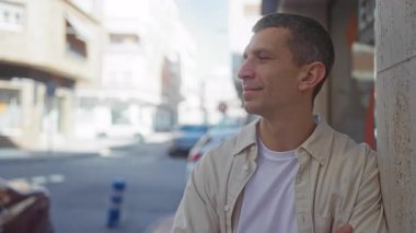 Güneşli bir sokakta güvenli bir şekilde gülümseyen bir adam. Açık havada park etmiş arabaları olan, binaların önünde rahat bir duruş takınan bir adam..