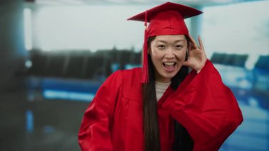 Havaalanı terminalinde kırmızı mezuniyet elbisesi içinde gülümseyen bir kadın baş parmağıyla kulaktan kulağa dinliyor..