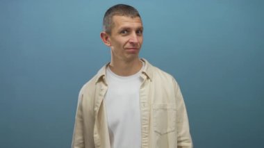 Kendine güvenen beyaz bir adam mavi izole bir arka plana gülümsüyor, bej bir gömlek ve beyaz bir tişört giyiyor, pozitif bir tavır sergiliyor..