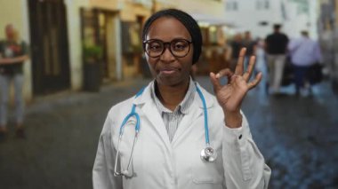 Sokakta steteskopla gülümseyen doktor kadın arka planda bulanık binalarla el hareketi gösteriyor..