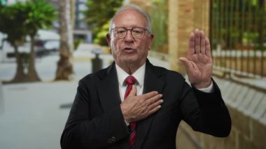 Takım elbiseli kıdemli bir adam açık havada, eli yüreğinde ve diğer eli havada, bir şehir caddesinde samimiyet yayıyor, profesyonelliği ve dürüstlüğü öneriyor..