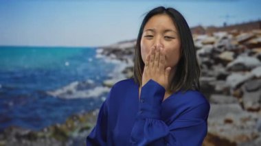 Mavi elbiseli genç bir kadın, okyanusun kenarında kaygısız bir anı yakalayan neşeli bir ifadeyle deniz kenarında öpücük yolluyor..