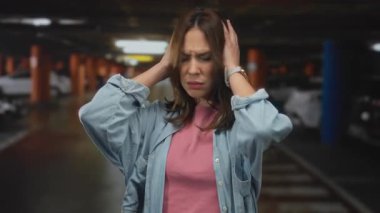 Kadın, kapalı otoparkta kot pantolon ve pembe gömlek giyerek hayal kırıklığına uğruyor. Arka planda loş bir ışık var. Günlük kentsel çevrede stres ve kafa karışıklığını temsil ediyor..