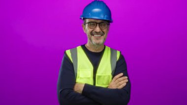 Kasketi ve yeleği olan kendine güvenen, kolları çapraz bir şekilde gülümseyen, canlı pembe arka planda inşaat endüstrisi ve güvenlik ekipmanlarını gösteren kendine güvenen bir adam..