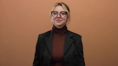 Kahverengi bir arkaplanda kollarını kavuşturup gülümseyen bir kadın. Kendine güvenen ve iş kadını gibi bir poz veren bir kadın. Gözlük takıyor ve profesyonel bir tavır sergileyen siyah bir ceket giyiyor..