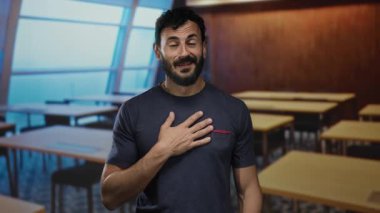 Göğsüne dokunarak şükranlarını ifade eden sakallı İspanyol bir adam, arka planda masalarla bir okul odasında duruyor..