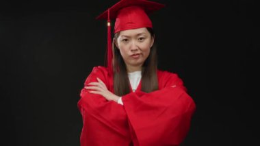 Ciddi bir ifadeyle mezuniyet elbisesi giymiş Asyalı bir kadın akademik başarı ve duyguyu vurgulayan siyah bir arkaplana karşı duruyor..