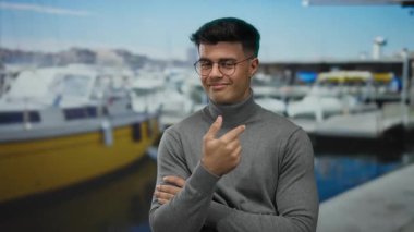 Gözlüklü genç adam deniz kıyısında arka planda tekneleri olan, gri balıkçı yakalı, açık havada kendine güvenen, onaylamadığını belirten, parmağını kaldırarak işaret eden bir jest yapıyor..
