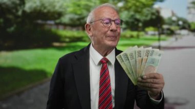 Parkta gülümseyen kıdemli işadamı elinde Czech Koruna banknotları, resmi bir ortamda finansal başarı ve mutluluğu somutlaştırıyor..