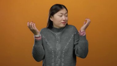 Stüdyo ortamında turuncu arka plan ile izole edilmiş süveter giyen, omuz silken bir kadın belirsizliği ya da sorgulama tutumunu öne sürüyor..