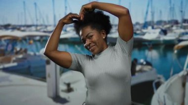 Gülümseyen Afrikalı Amerikalı kadın, güneşli bir açık havada, deniz kıyısında teknelerle el kol hareketi yapıyor, neşe ve coşku dolu atmosfer sergiliyor..
