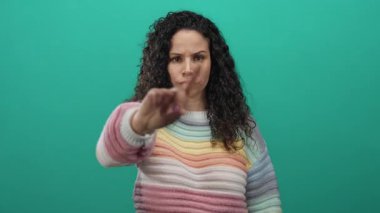 Yeşil arka planda kameraya doğru elini kaldıran, renkli kazak giyen, reddedilmeyi ciddi bir ifadeyle ifade eden bir kadın..