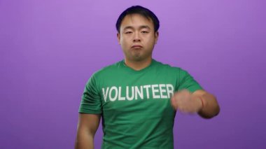 Yeşil gönüllü tişörtü giymiş, kapalı gözlerle selam veren genç adam izole edilmiş mor arka plana karşı duruyor..