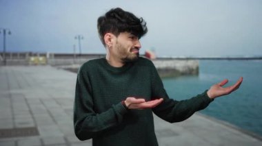 Kafası karışmış genç bir adam okyanus manzaralı deniz kenarı gezinti alanında dikiliyor. Koyu renkli bir süveter giyiyor. Açık hava kentsel davranışları var.