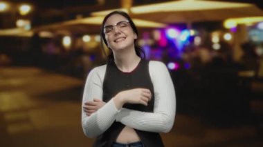 Gece canlı bir terasta gözlüklü gülümseyen bir kadın. Bulanık restoran ışıklarıyla sıcak bir sokak havası yaratıyor..