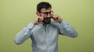 Sakallı ve gözlüklü İspanyol bir adam, sarı arka plandaki üzüntüyü taklit eder. Duygularını yüzüne yakın ifade eder, mavi gömlek giyer..