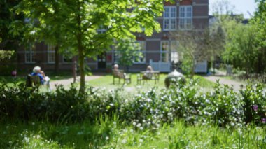 Yeşilliklerle çevrili banklarda oturan insanların oturduğu bulanık park manzarası ve tarihi bir yapı zemini. Doğanın dokunuşuyla huzurlu ve rahat bir atmosfer yaratıyor..