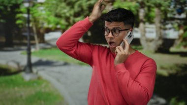Kırmızı kazaklı genç adam yeşil park manzarasıyla çevrili bir akıllı telefondan konuşuyor..