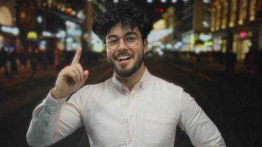 Gözlüklü, sakallı, şehir ışıklarıyla aydınlatılmış genç bir İspanyol gece açık havayı işaret ediyor. Bu da kalabalık bir şehir ortamında keşfedildiğini gösteriyor..