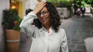 Beyaz tişörtlü, kıvırcık saçlı, şehir sokaklarında düşünceli görünen bir kadın..