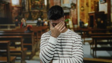 Çizgili gömlekli genç bir adam kilisenin içinde elleri yüzünde düşünceli görünüyor, huzurlu bir atmosferde boş sıralarla çevrili..