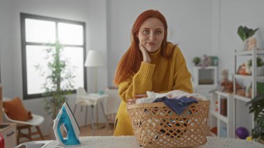 Kızıl saçlı kadın çamaşırlarını çamaşır odasında hasır sepet ve demirle temizliyor.