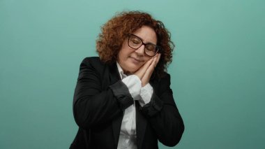 Siyah takım elbiseli, kıvırcık esmer saçlı orta yaşlı bir kadın yeşil bir duvar arkasında izole edilmiş bir şekilde uyku jesti yapıyor..